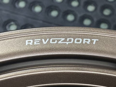Bronze REVOZPORT forged alloy wheel with an intricate split multi-spoke design displayed on a black textured workshop floor.
