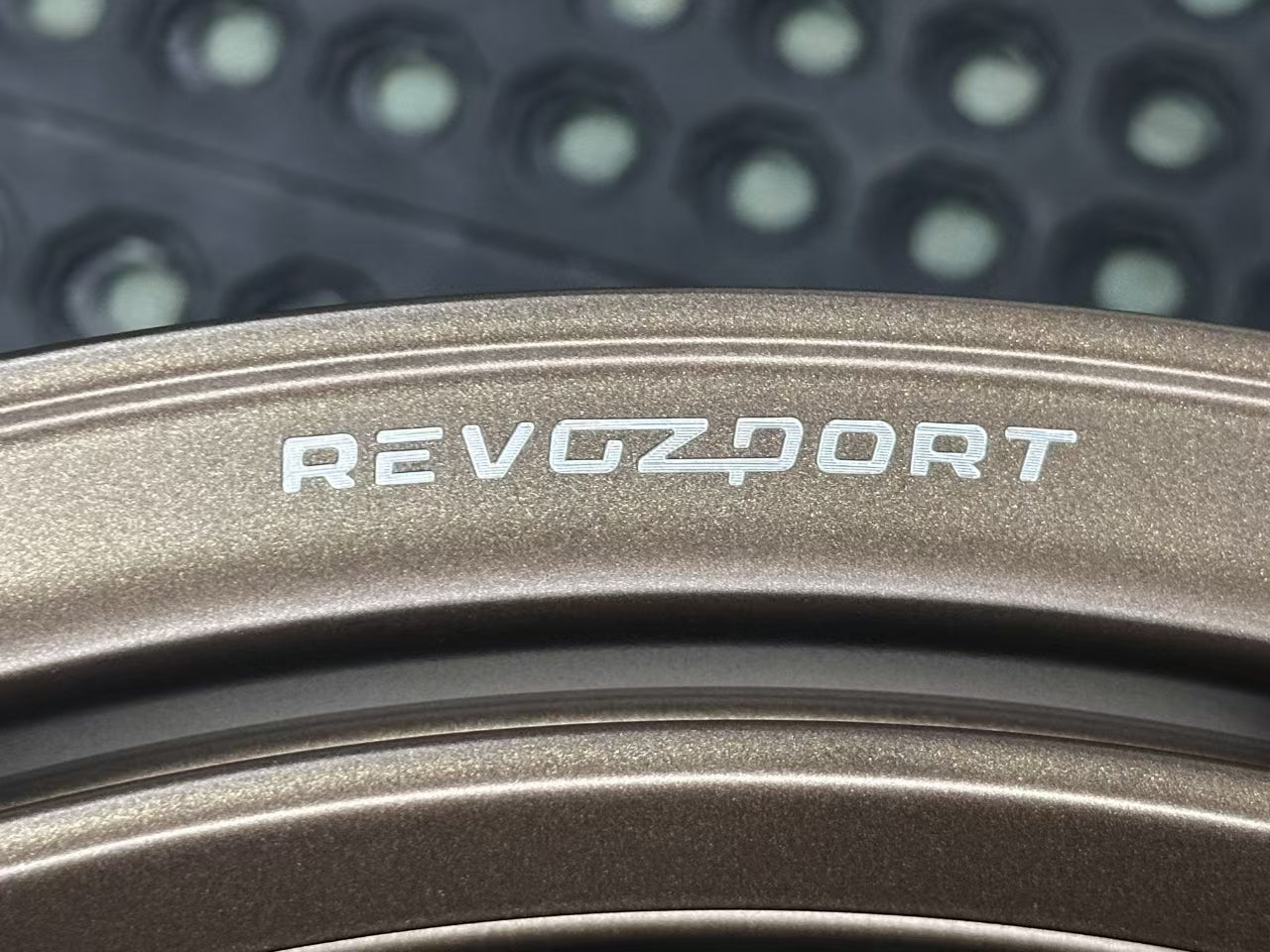 Bronze REVOZPORT forged alloy wheel with an intricate split multi-spoke design displayed on a black textured workshop floor.