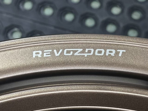 Bronze REVOZPORT forged alloy wheel with an intricate split multi-spoke design displayed on a black textured workshop floor.
