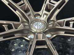 Bronze REVOZPORT forged alloy wheel with an intricate split multi-spoke design displayed on a black textured workshop floor.