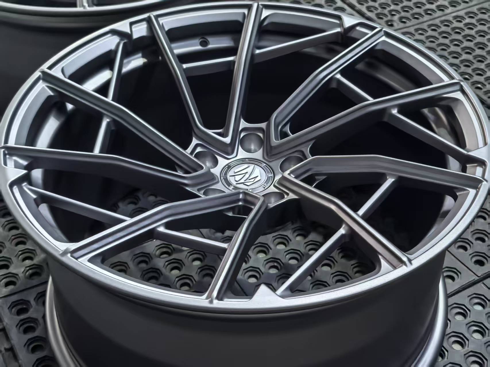 Matte gray REVOZPORT forged alloy wheel with an intricate split multi-spoke design displayed on a black textured workshop floor.