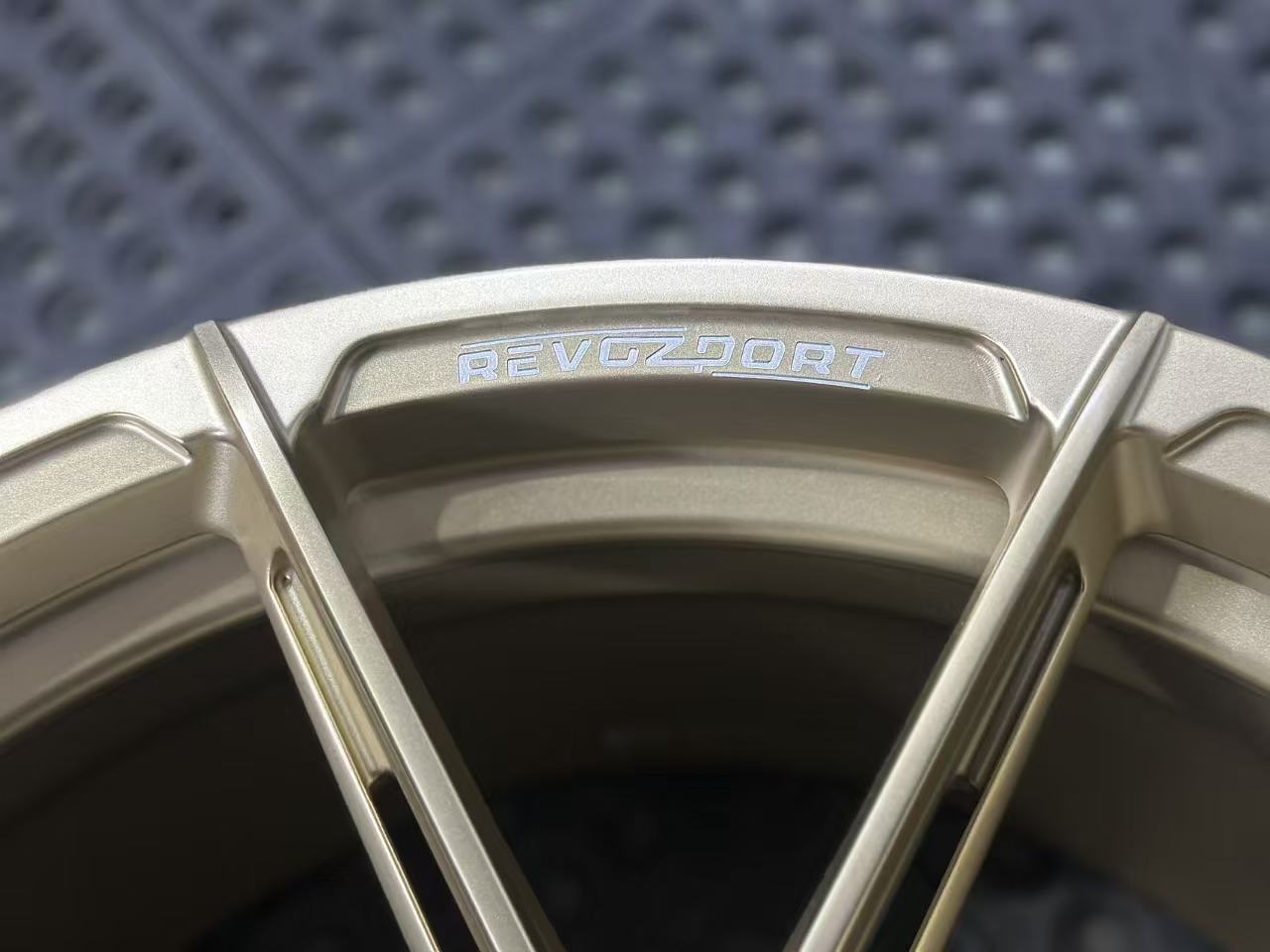 Bronze REVOZPORT forged alloy wheel with split multi-spoke design displayed on a black textured workshop floor.