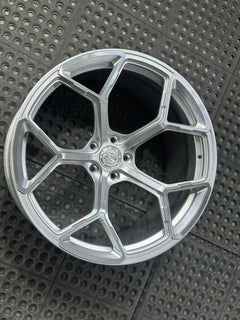 Silver forged alloy wheel with a multi-spoke design displayed against a black textured rubber floor.