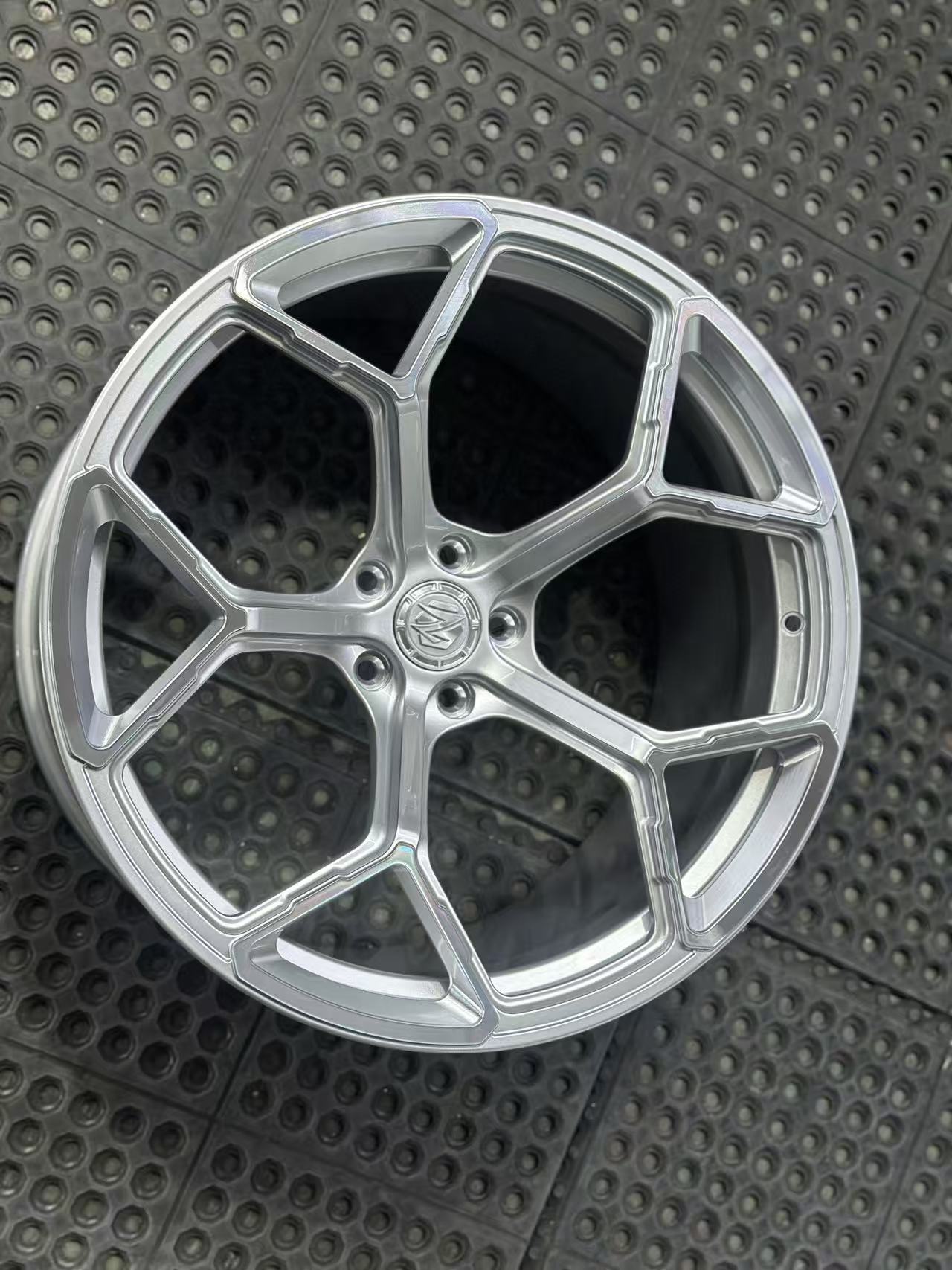 Silver forged alloy wheel with a multi-spoke design displayed against a black textured rubber floor.