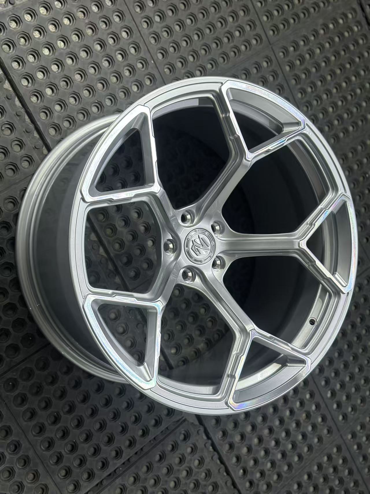 Silver forged alloy wheel with a multi-spoke design displayed against a black textured rubber floor.