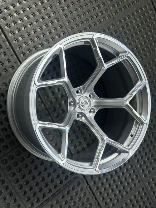Silver forged alloy wheel with a multi-spoke design displayed against a black textured rubber floor.