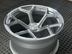 Silver forged alloy wheel with a multi-spoke design displayed against a black textured rubber floor.