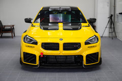 Yellow BMW G87 M2 featuring a REVOZPORT carbon fiber body kit with vented hood, front splitter, large air intakes, and aggressive track-focused aero styling displayed indoors.