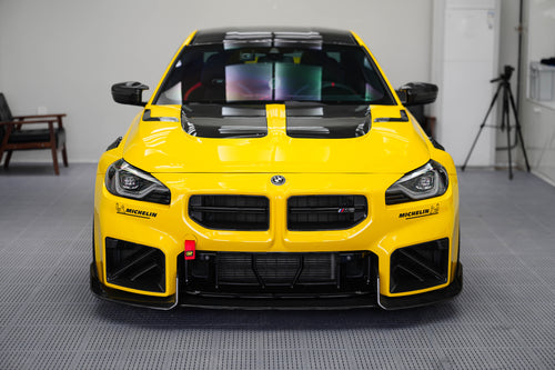 Yellow BMW G87 M2 featuring a REVOZPORT carbon fiber body kit with vented hood, front splitter, large air intakes, and aggressive track-focused aero styling displayed indoors.