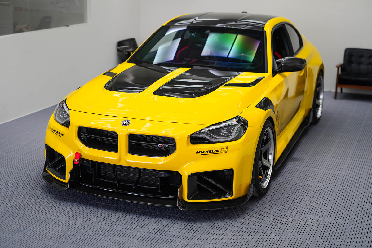 Yellow BMW G87 M2 featuring a REVOZPORT carbon fiber body kit with vented hood, front splitter, large air intakes, and aggressive track-focused aero styling displayed indoors.