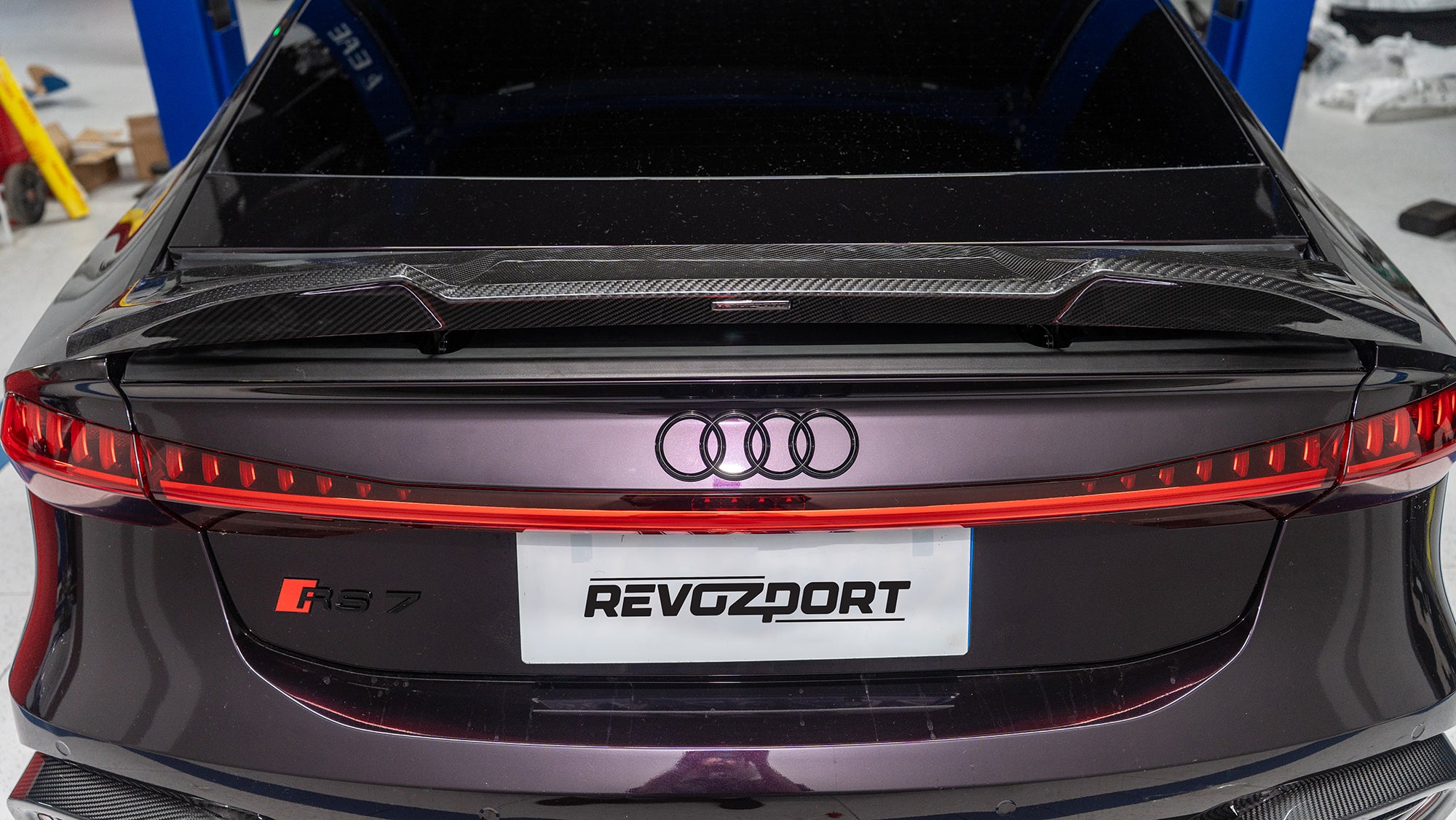 Black Audi RS7 featuring a REVOZPORT carbon fiber body kit with front lip, side skirts, carbon hood, and aggressive aero styling driving on a city street.