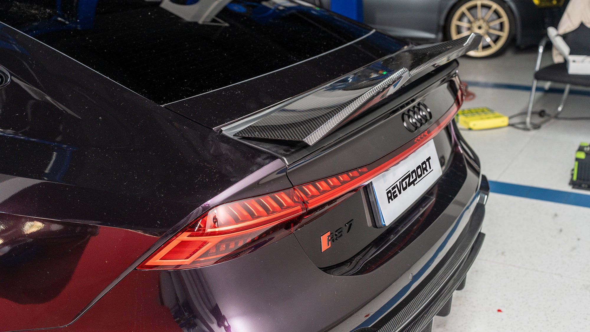 Black Audi RS7 featuring a REVOZPORT carbon fiber body kit with front lip, side skirts, carbon hood, and aggressive aero styling driving on a city street.