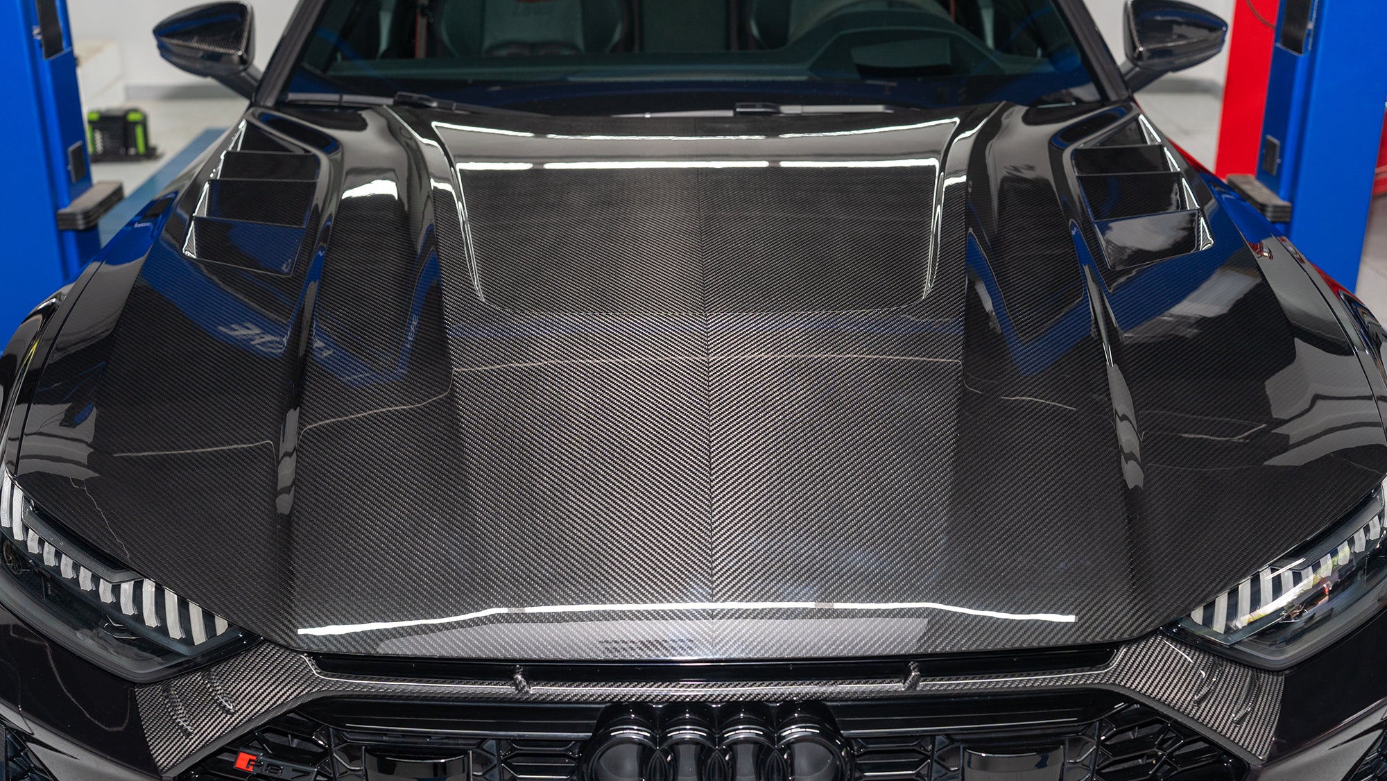 Black Audi RS7 featuring a REVOZPORT carbon fiber body kit with front lip, side skirts, carbon hood, and aggressive aero styling driving on a city street.