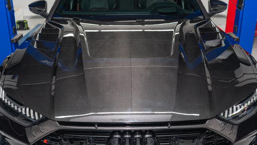 Black Audi RS7 featuring a REVOZPORT carbon fiber body kit with front lip, side skirts, carbon hood, and aggressive aero styling driving on a city street.