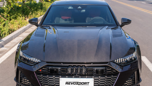 Black Audi RS7 featuring a REVOZPORT carbon fiber body kit with front lip, side skirts, carbon hood, and aggressive aero styling driving on a city street.