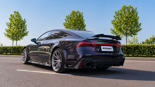 Black Audi RS7 featuring a REVOZPORT carbon fiber body kit with front lip, side skirts, carbon hood, and aggressive aero styling driving on a city street.