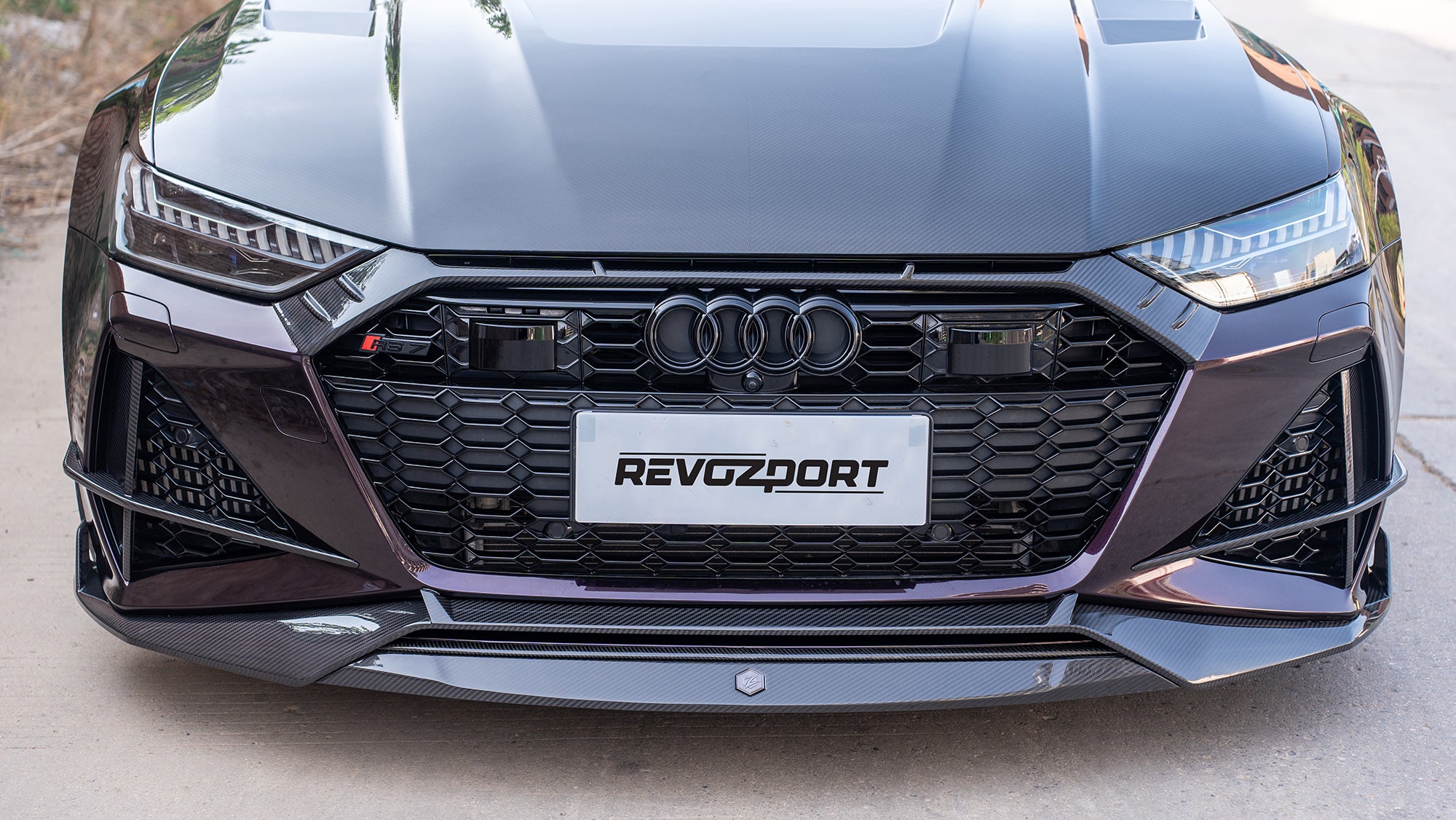 Black Audi RS7 featuring a REVOZPORT carbon fiber body kit with front lip, side skirts, carbon hood, and aggressive aero styling driving on a city street.