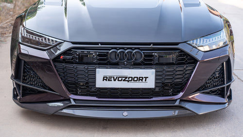 Black Audi RS7 featuring a REVOZPORT carbon fiber body kit with front lip, side skirts, carbon hood, and aggressive aero styling driving on a city street.