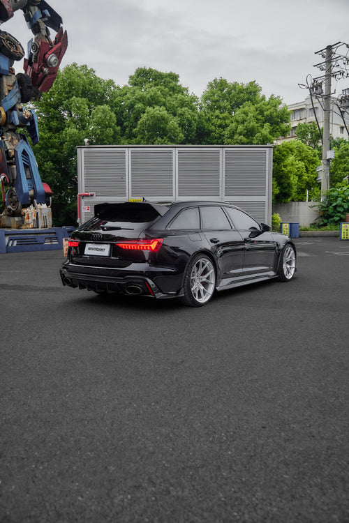 Black Audi RS6 Avant fitted with a REVOZPORT body kit and silver multi-spoke wheels, parked in an open lot surrounded by trees.