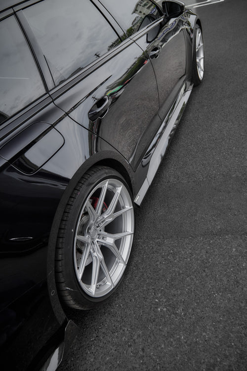 Black Audi RS6 Avant fitted with a REVOZPORT body kit and silver multi-spoke wheels, parked in an open lot surrounded by trees.