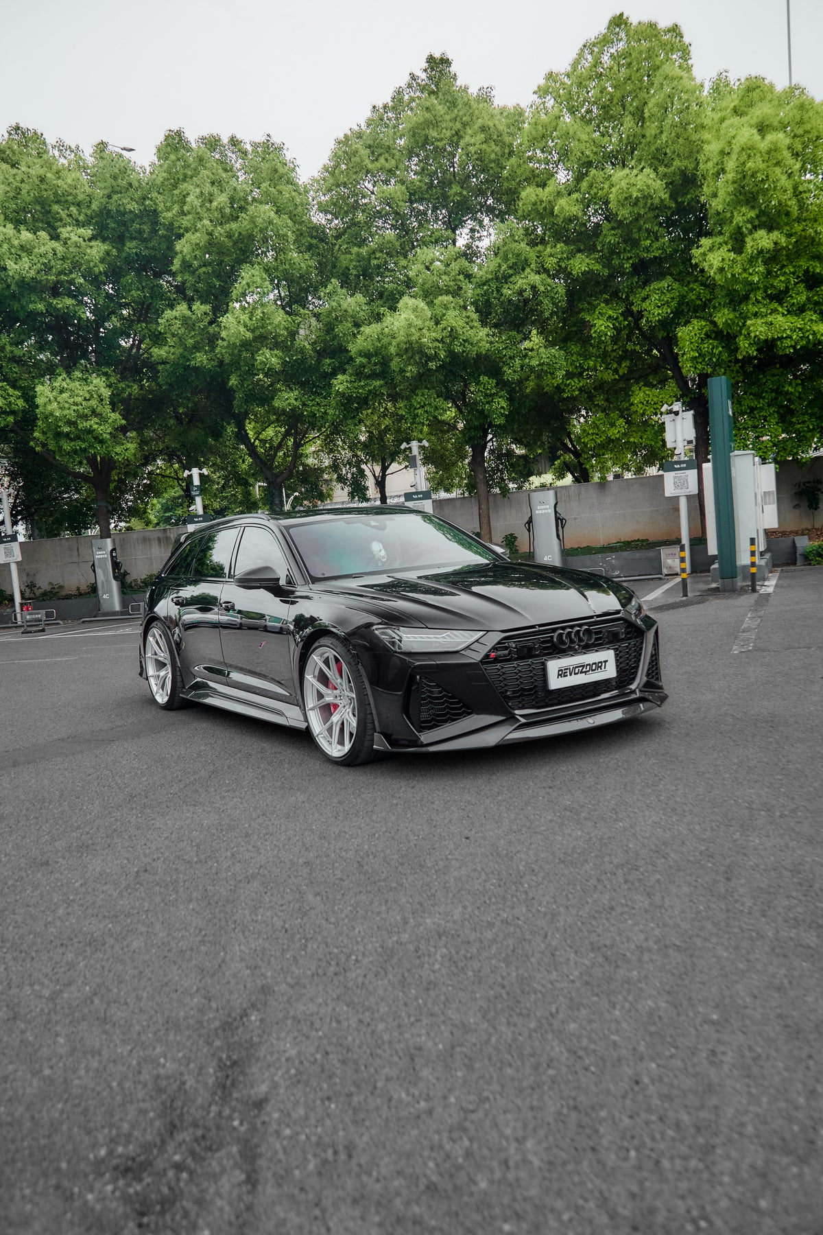 Black Audi RS6 Avant fitted with a REVOZPORT body kit and silver multi-spoke wheels, parked in an open lot surrounded by trees.