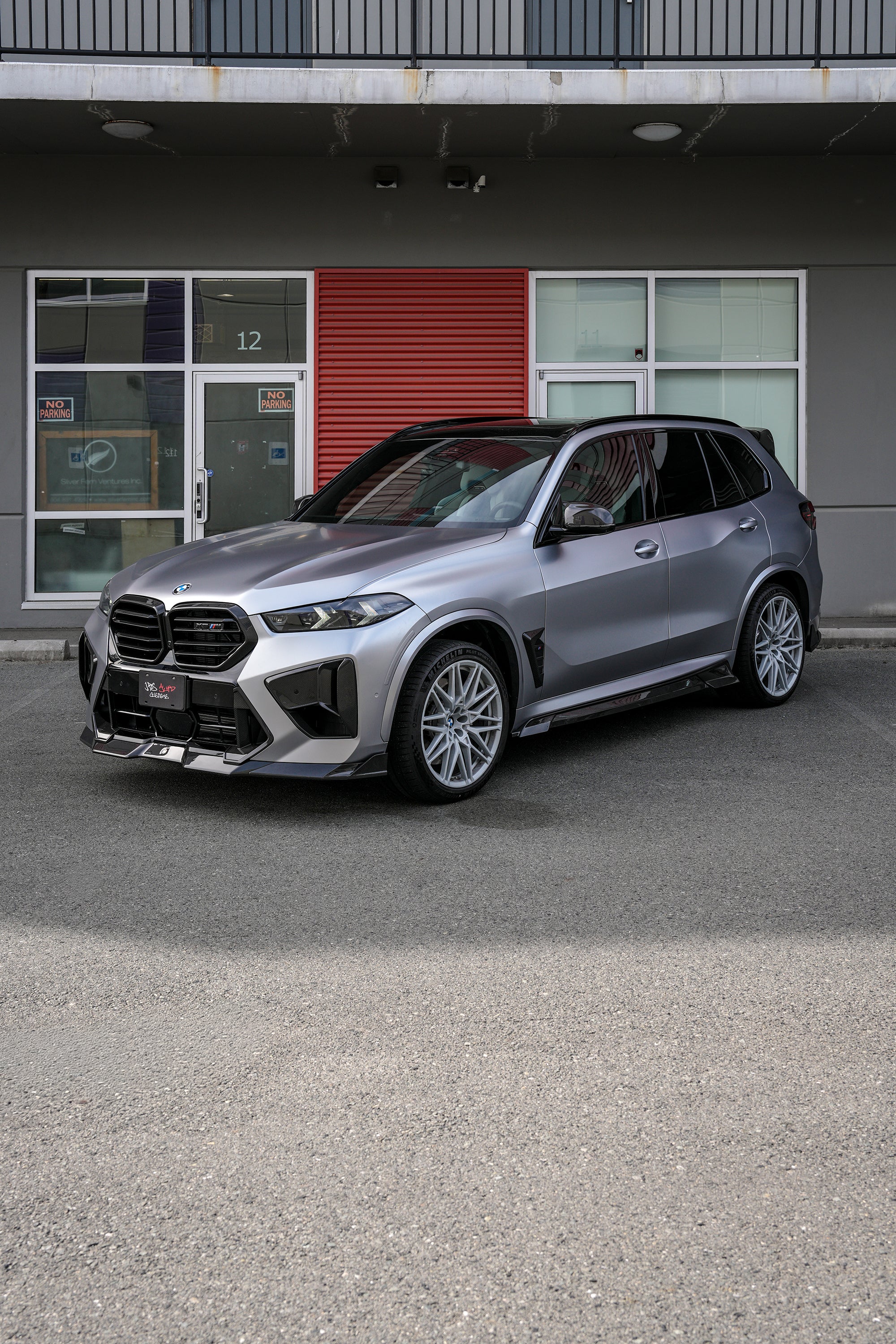 Gray BMW X5M featuring a REVOZPORT carbon fiber body kit with aggressive front lip, side skirts, and sporty aero styling parked outside a workshop.