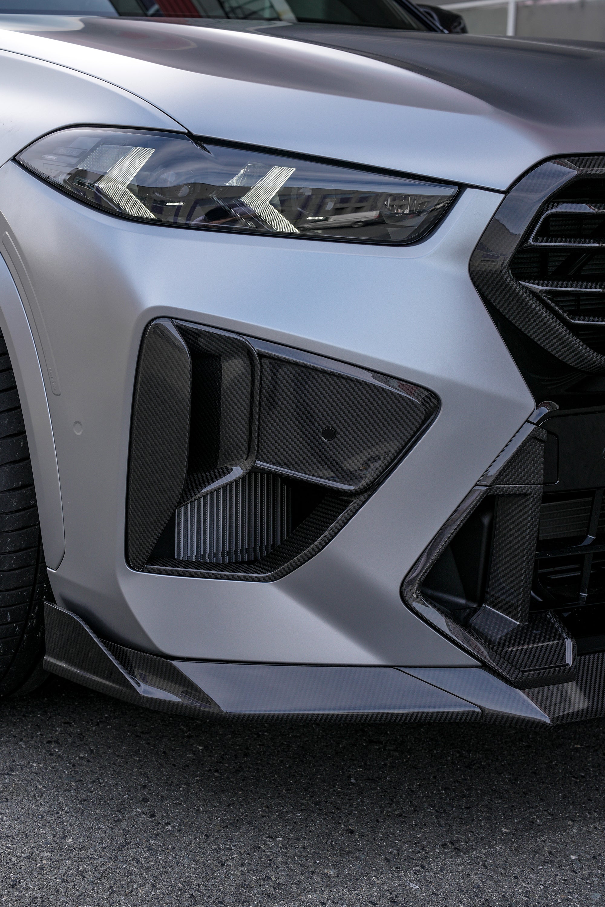 Gray BMW X5M featuring a REVOZPORT carbon fiber body kit with aggressive front lip, side skirts, and sporty aero styling parked outside a workshop.