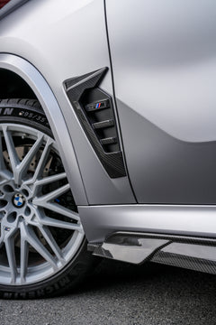 Gray BMW X5M featuring a REVOZPORT carbon fiber body kit with aggressive front lip, side skirts, and sporty aero styling parked outside a workshop.