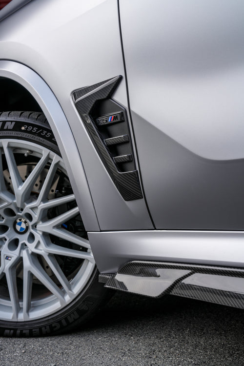 Gray BMW X5M featuring a REVOZPORT carbon fiber body kit with aggressive front lip, side skirts, and sporty aero styling parked outside a workshop.