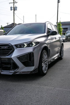 Gray BMW X5M featuring a REVOZPORT carbon fiber body kit with aggressive front lip, side skirts, and sporty aero styling parked outside a workshop.