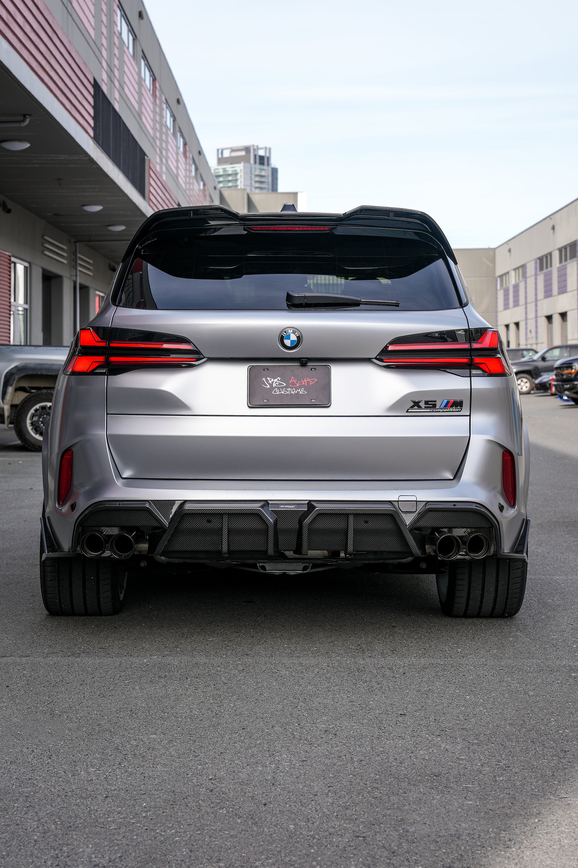 Gray BMW X5M featuring a REVOZPORT carbon fiber body kit with aggressive front lip, side skirts, and sporty aero styling parked outside a workshop.