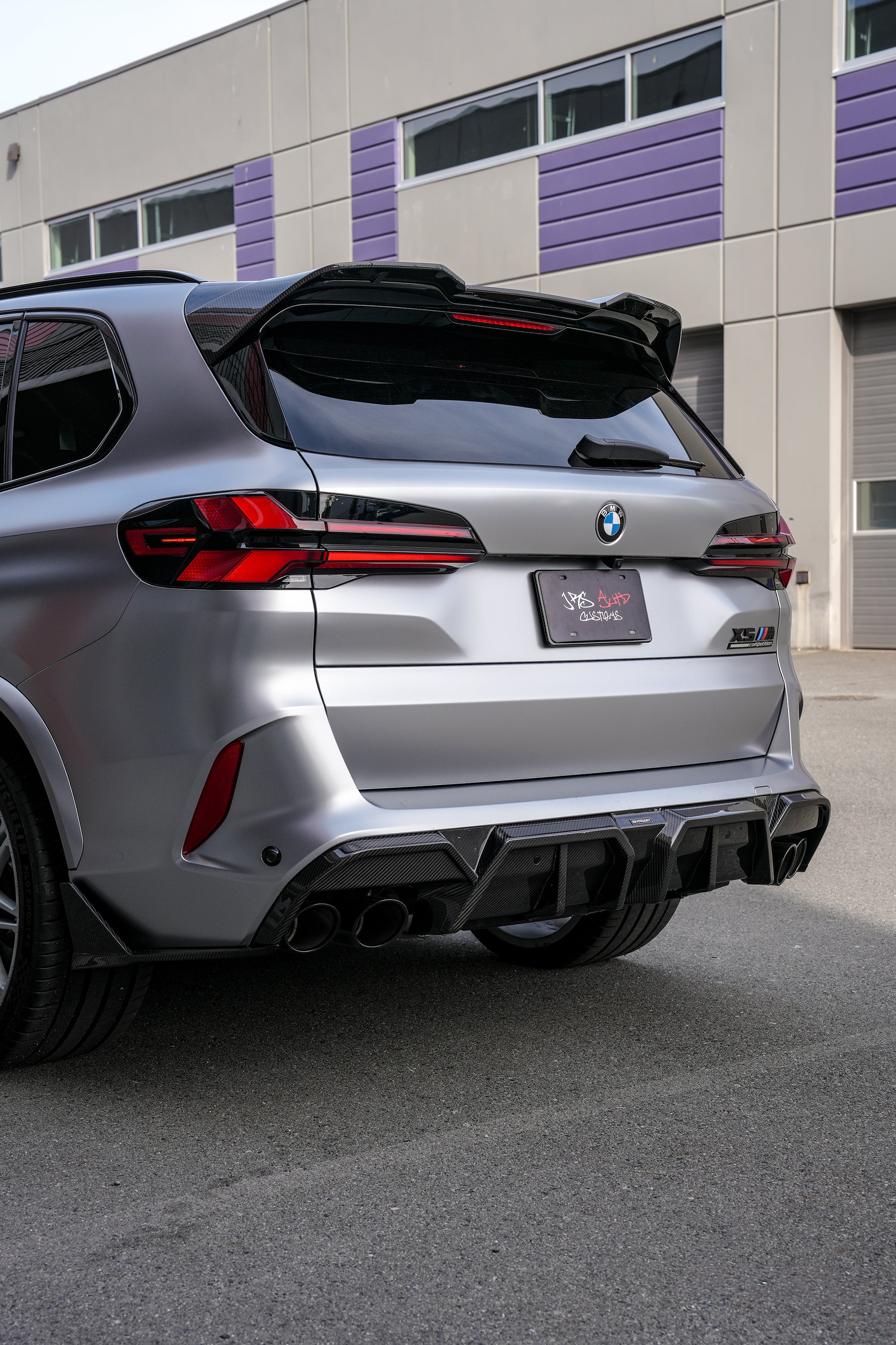 Gray BMW X5M featuring a REVOZPORT carbon fiber body kit with aggressive front lip, side skirts, and sporty aero styling parked outside a workshop.