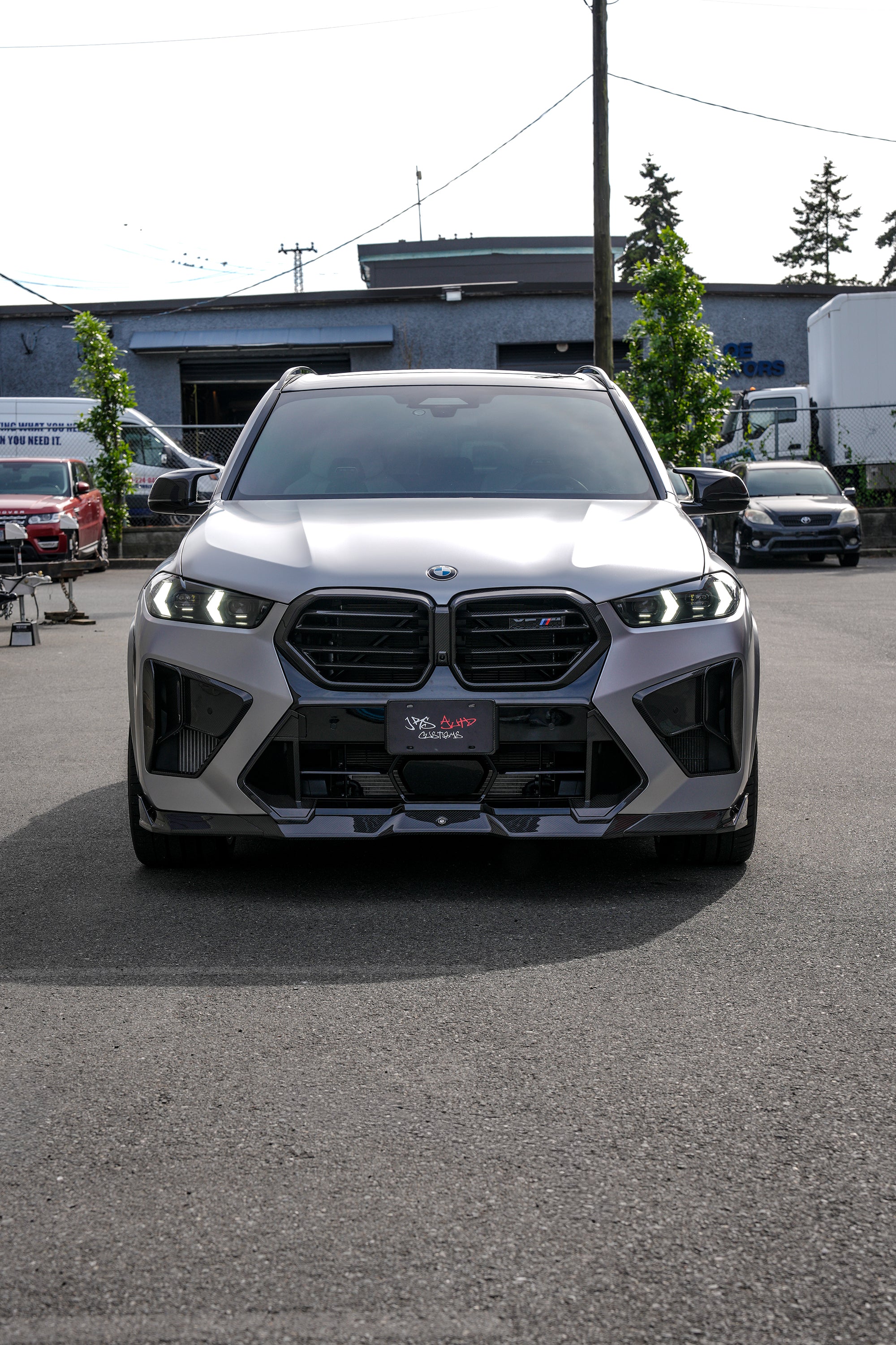 Gray BMW X5M featuring a REVOZPORT carbon fiber body kit with aggressive front lip, side skirts, and sporty aero styling parked outside a workshop.