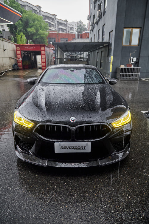 Rain-covered BMW front view with yellow headlights, carbon fiber hood, front lip, and aggressive aero styling on wet pavement.