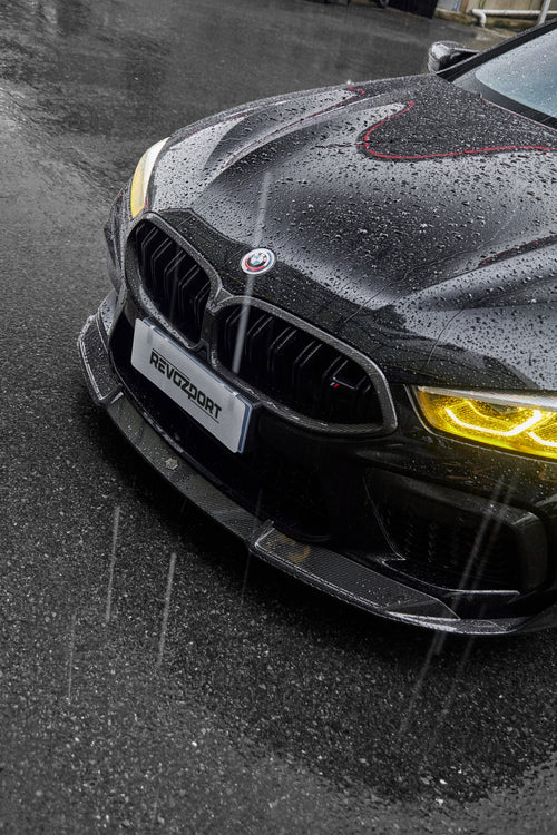 Rain-covered BMW front close-up with yellow headlights, carbon fiber hood, front lip, and aggressive aero styling.