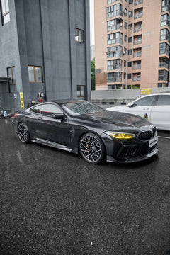 Rain-covered BMW with carbon fiber body kit, yellow headlights, and aggressive stance parked on wet pavement in an urban setting.