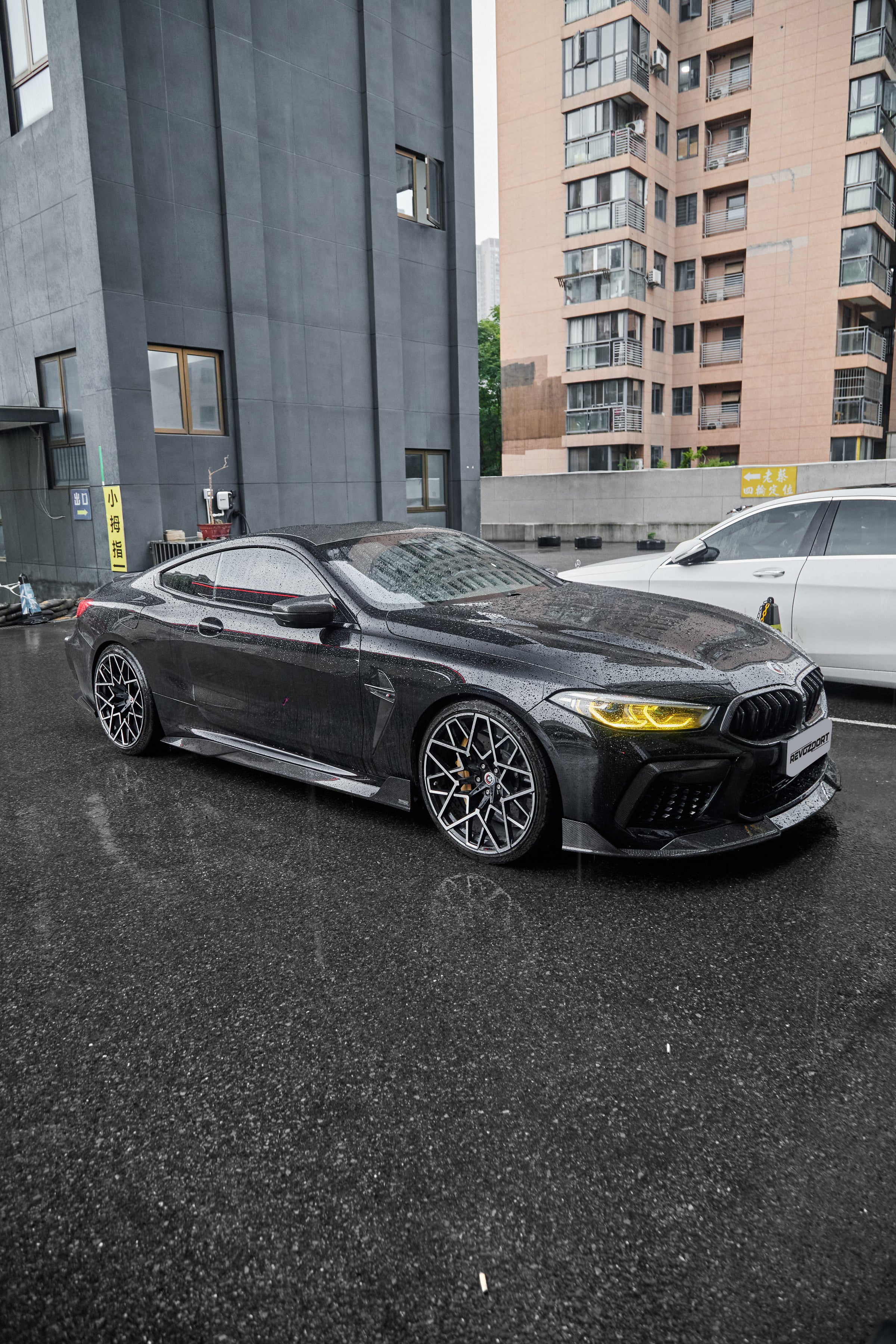 Rain-covered BMW with carbon fiber body kit, yellow headlights, and aggressive stance parked on wet pavement in an urban setting.