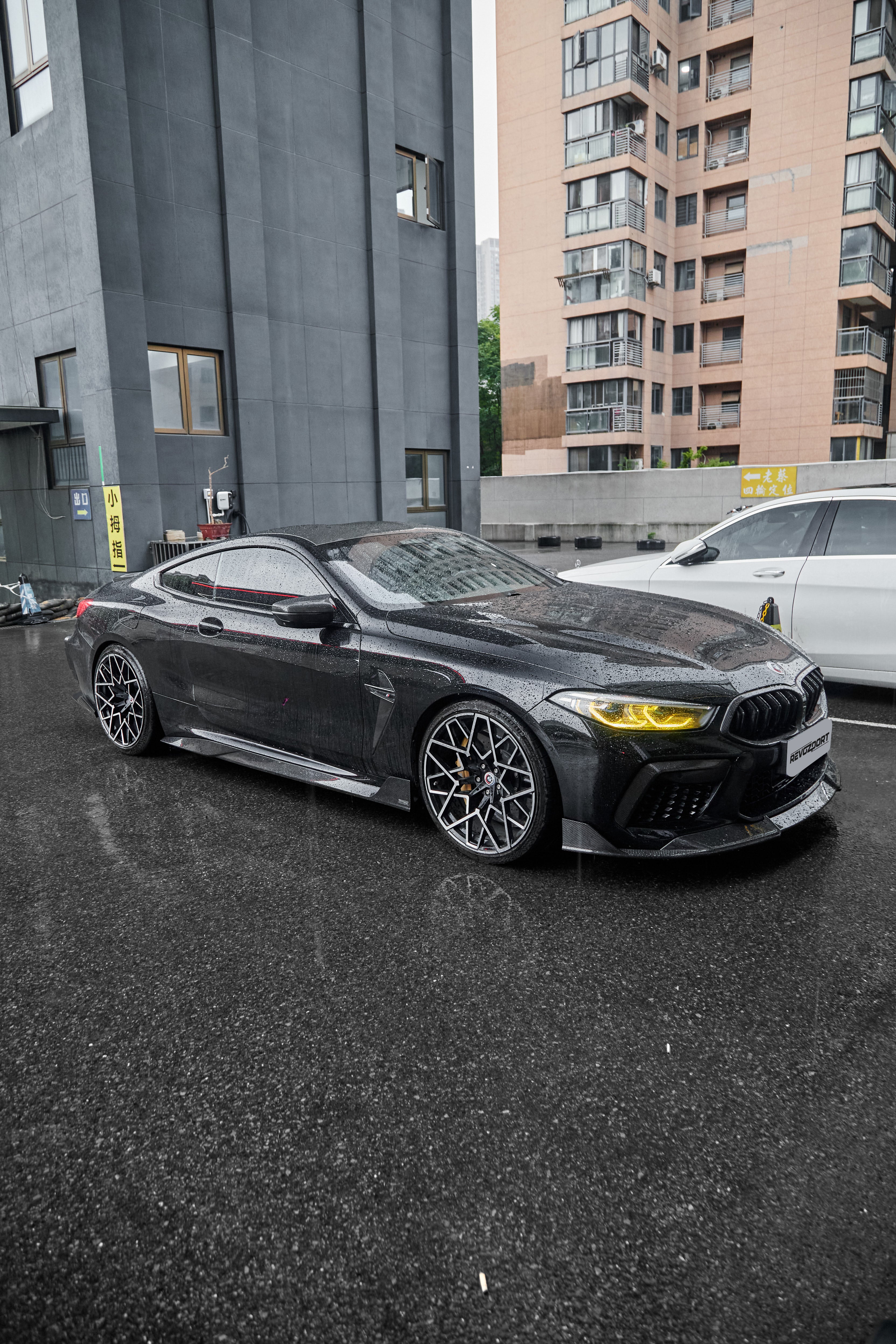 Rain-covered BMW with carbon fiber body kit, yellow headlights, and aggressive stance parked on wet pavement in an urban setting.