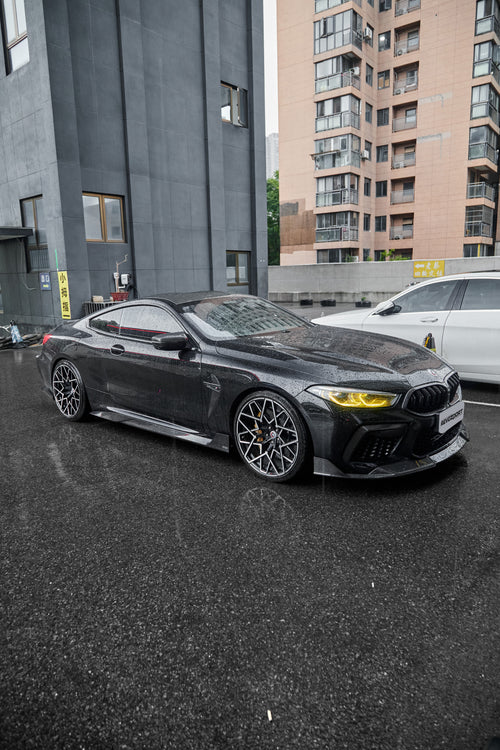 Rain-covered BMW with carbon fiber body kit, yellow headlights, and aggressive stance parked on wet pavement in an urban setting.