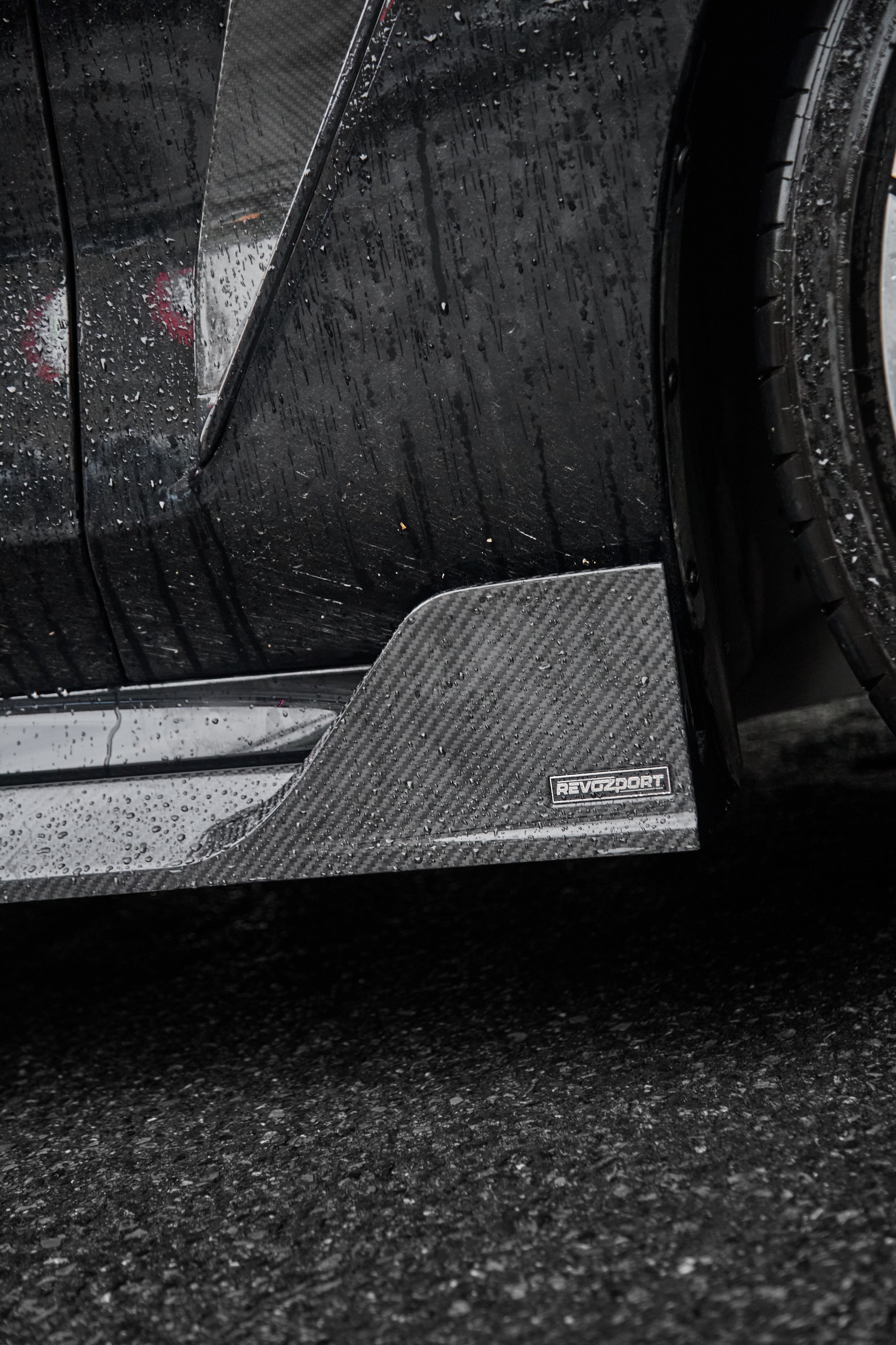 Close-up of a rain-covered carbon fiber side skirt and aero fin, highlighting glossy weave texture and precision detailing.