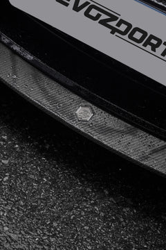 Close-up of a rain-covered carbon fiber front lip, highlighting glossy weave texture, precision badge detail, and wet pavement.