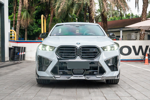 Front-facing view of a white BMW featuring aggressive carbon fiber front aero, illuminated headlights, sharp intake details, and performance-focused styling.