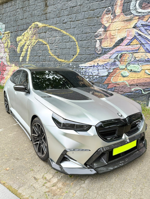 Silver BMW with carbon fiber body kit parked beside a colorful graffiti wall, highlighting front lip, hood vents, and aggressive styling.