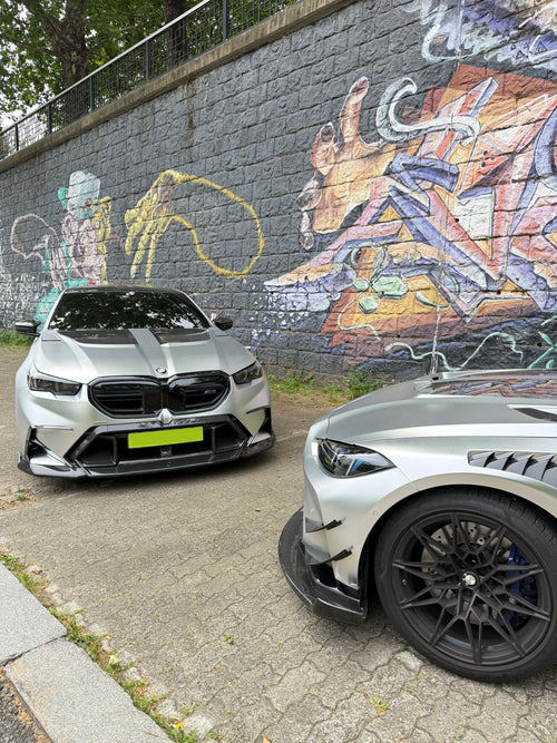 Two BMWs with carbon fiber aero kits parked beside a colorful graffiti wall, highlighting aggressive front styling and urban atmosphere.