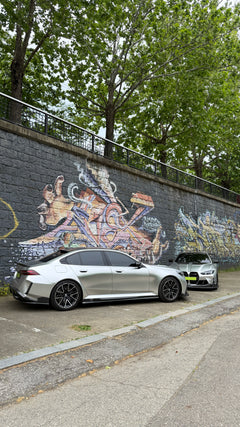 Top-down view of two BMWs with carbon fiber aero kits, hood vents, and rear wing parked beside a quiet road.