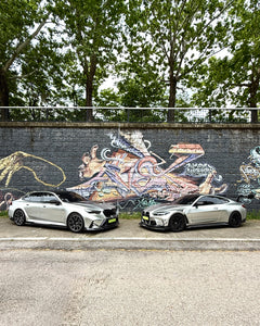 Two BMWs with carbon fiber aero kits parked side by side against a colorful graffiti wall in an urban street setting.