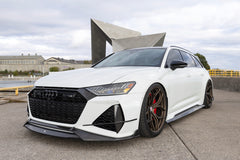 White Audi RS6 Avant featuring a REVOZPORT carbon fiber body kit with front lip, side skirts, rear diffuser, and lowered stance parked in a modern urban waterfront setting.