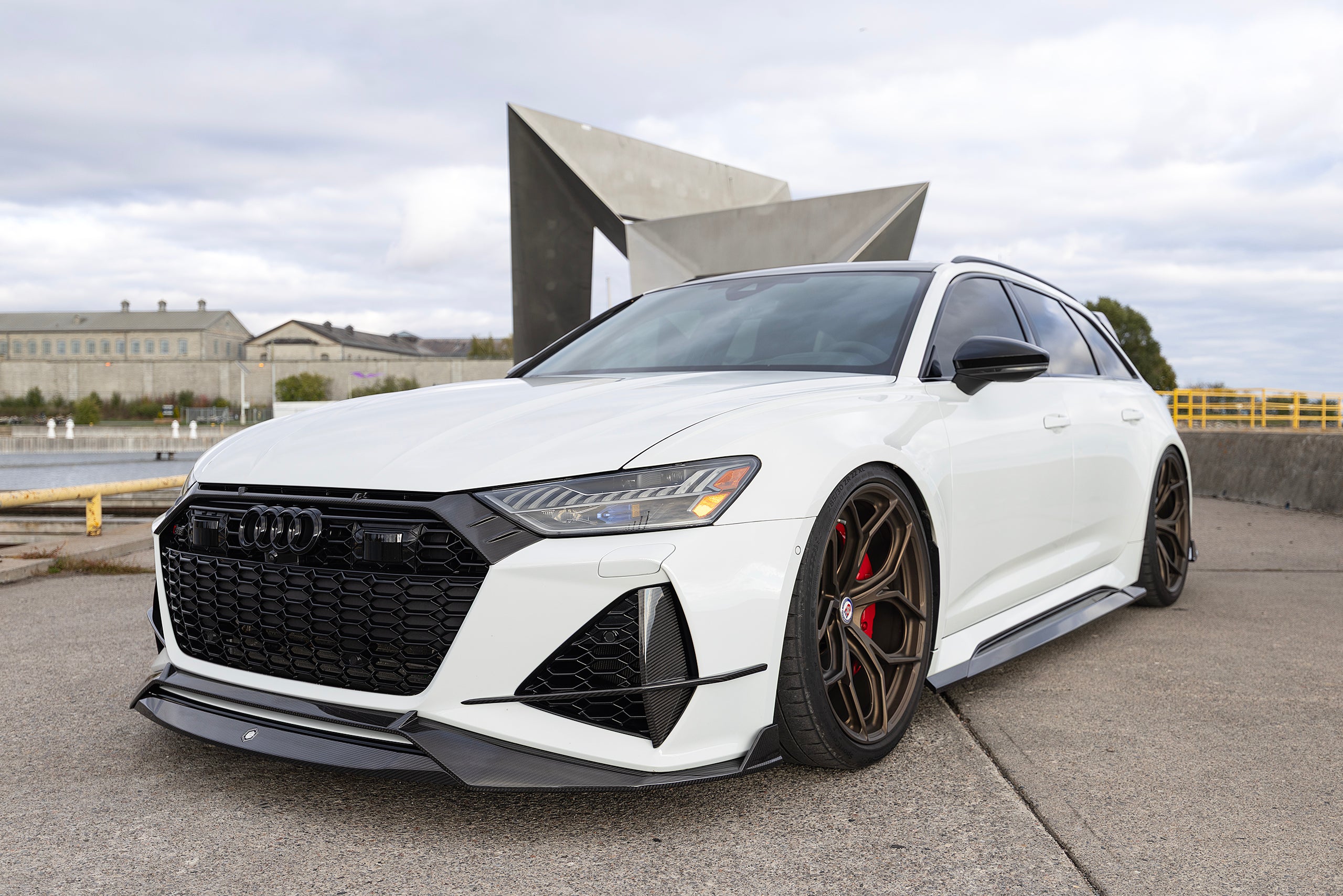 White Audi RS6 Avant featuring a REVOZPORT carbon fiber body kit with front lip, side skirts, rear diffuser, and lowered stance parked in a modern urban waterfront setting.