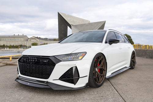 White Audi RS6 Avant featuring a REVOZPORT carbon fiber body kit with front lip, side skirts, rear diffuser, and lowered stance parked in a modern urban waterfront setting.