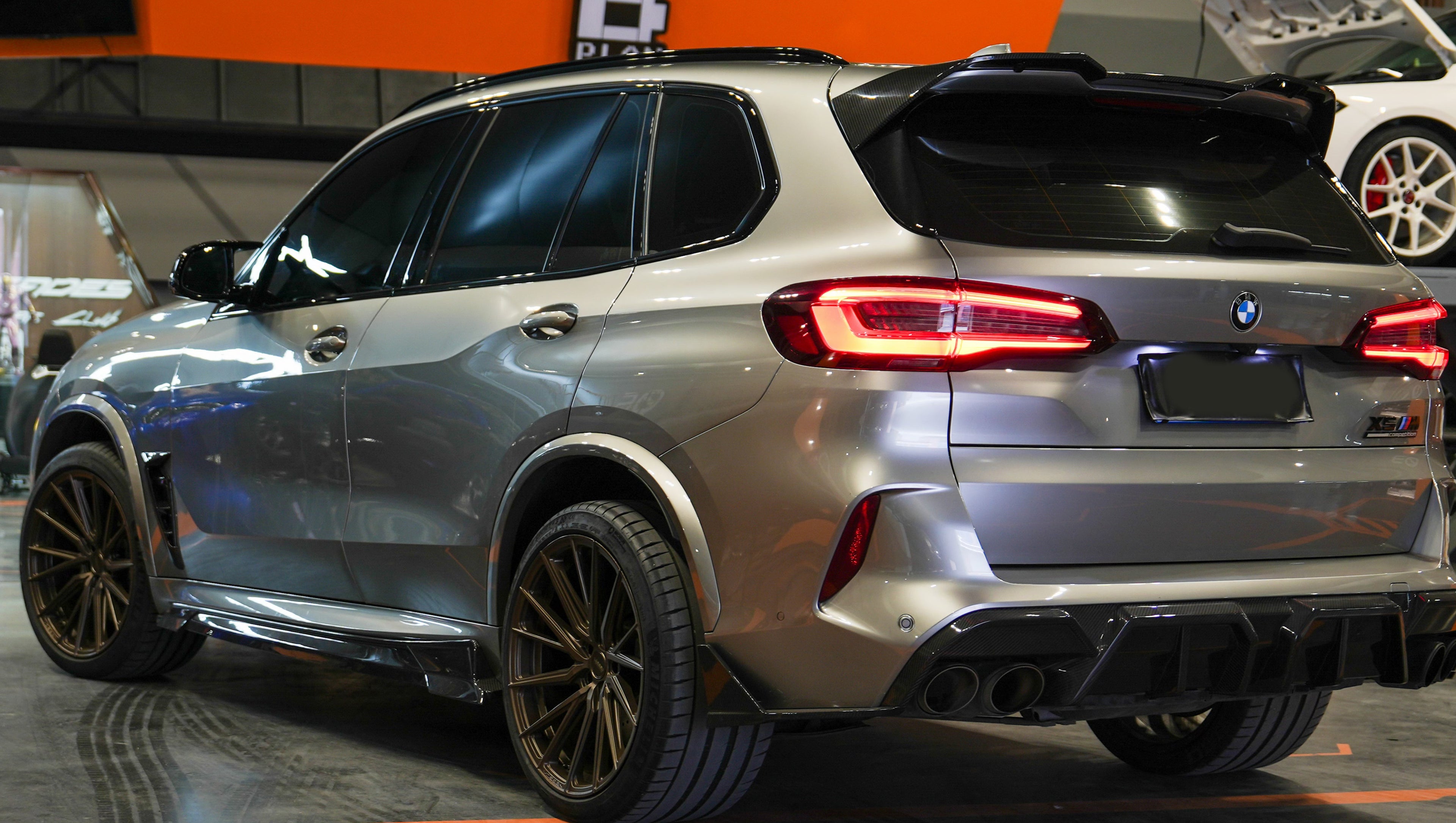 Gray BMW X5M featuring a REVOZPORT carbon fiber body kit with aggressive front lip, enlarged air intakes, side skirts, and sporty aero styling displayed inside a performance workshop.